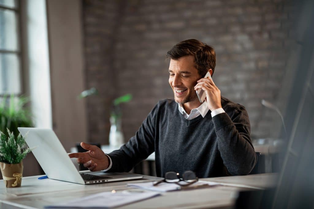 businessman talking on cell phone Happy businessman communicating over mobile phone and working on laptop while being at work.
