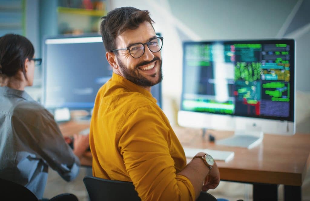 Closeup over the shoulder view of a cheerful mid 20's software developer at the office smiling to the camera. His female coworker is next him, still focused and working.