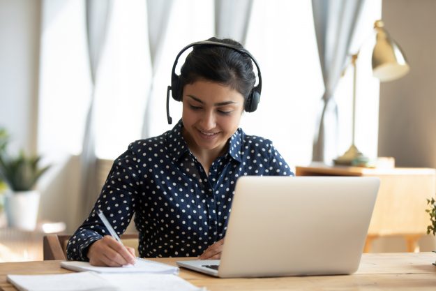 Happy indian girl student wear headset study online with webcam teacher write notes, happy young woman listen lecture watch webinar on laptop sit at desk, distance e learn language education concept