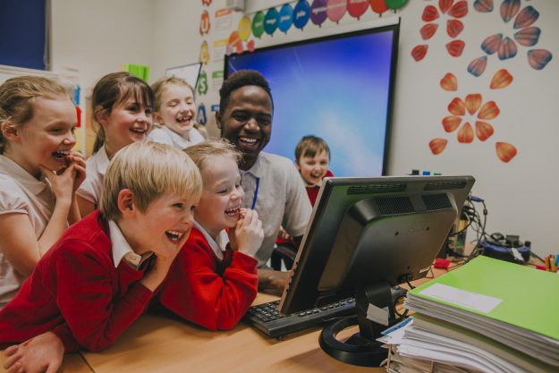 Group of primary school students are crowded round a computer with their teacher. They are all laughing and looking at the computer screen.